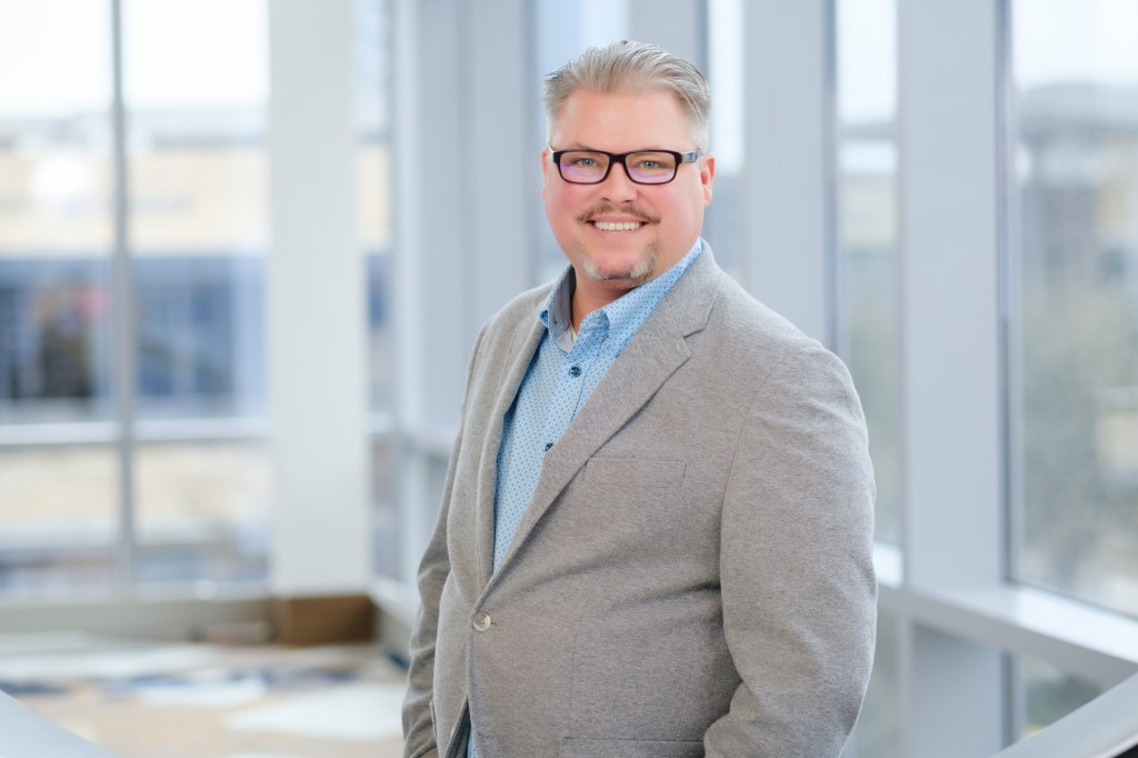 A smiling man with glasses stands confidently in front of large windows, dressed in a gray blazer over a blue shirt. omni dallas headshot photographer nicole caldwell