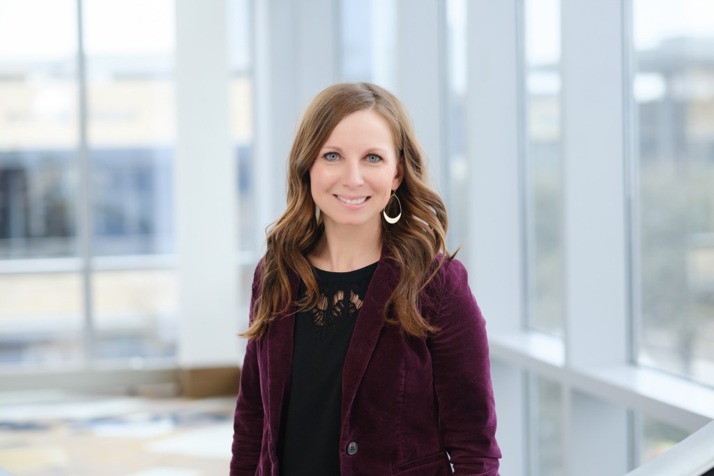 A professional headshot of a woman with long brown hair, wearing a dark top and a burgundy blazer, smiling in front of large windows in a bright indoor setting. omni dallas headshot photographer nicole caldwell