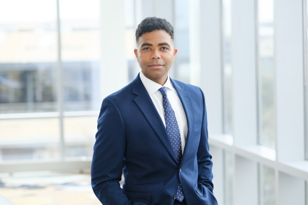 A professional portrait of a man in a blue suit with a white shirt and polka dot tie, standing confidently indoors against a backdrop of large windows. omni dallas headshot photographer nicole caldwell