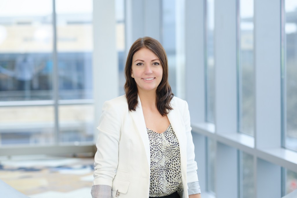 A woman with shoulder-length brown hair wearing a light-colored blazer and a patterned top smiles in a well-lit office with large windows. omni dallas headshot photographer nicole caldwell