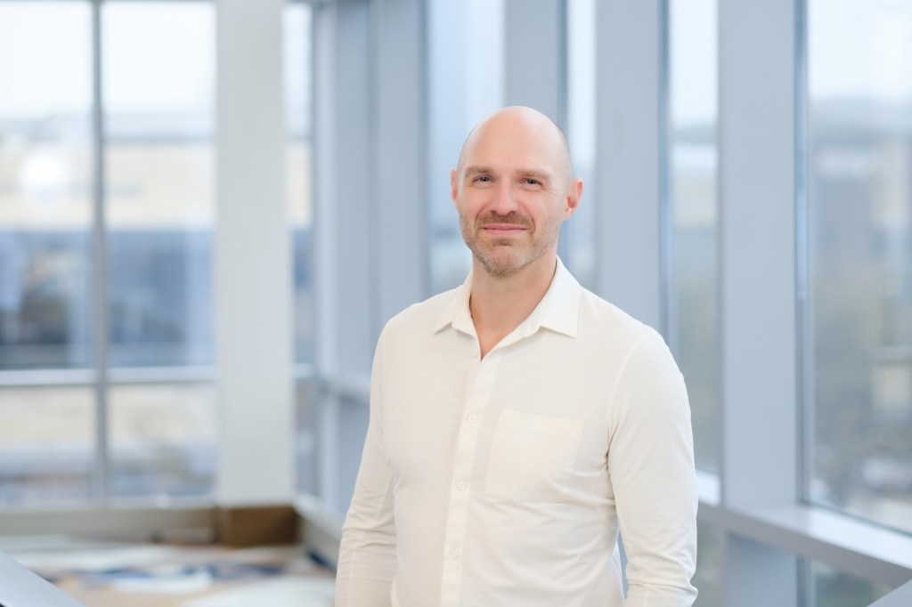 A smiling man wearing a white shirt stands indoors in front of large windows, with a modern office space visible in the background. omni dallas headshot photographer nicole caldwell