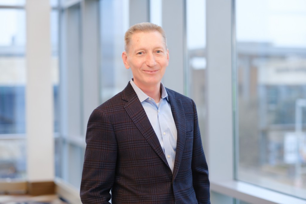 Professional headshot of a man smiling, wearing a dark blazer and light blue shirt, standing by a large window with a view of an urban setting. omni dallas headshot photographer nicole caldwell