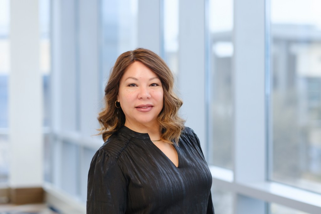 A woman with shoulder-length brown hair wearing a black top, smiling confidently in front of large windows at a seminar venue. omni dallas headshot photographer nicole caldwell