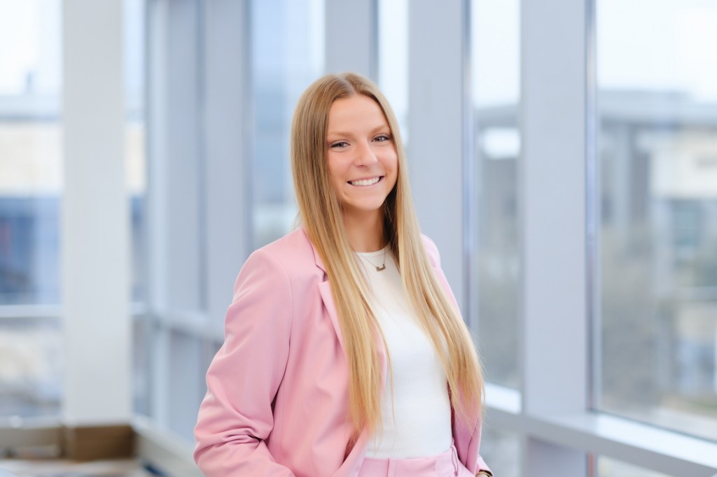 Smiling woman in a pink suit standing by large windows, showcasing a bright and modern indoor setting. omni dallas headshot photographer nicole caldwell