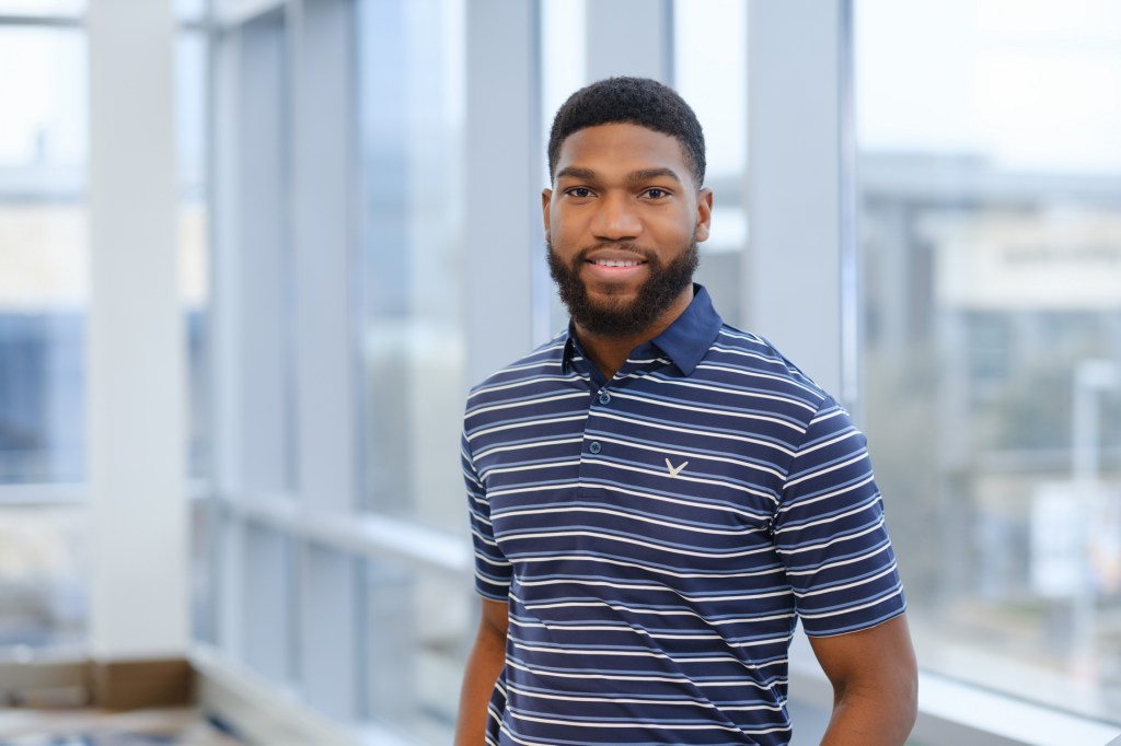 Smiling man with a beard wearing a striped polo shirt, posing indoors with large windows in the background. omni dallas headshot photographer nicole caldwell