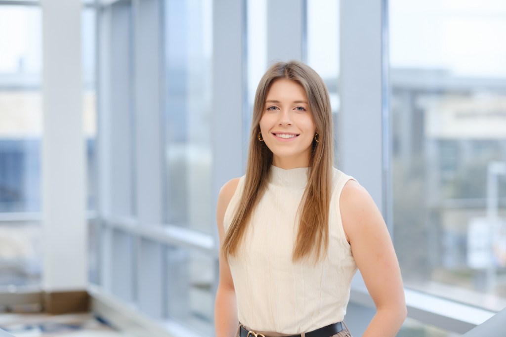 A smiling female portrait subject in a light-colored sleeveless top, standing near a large window with soft, natural light. omni dallas headshot photographer nicole caldwell