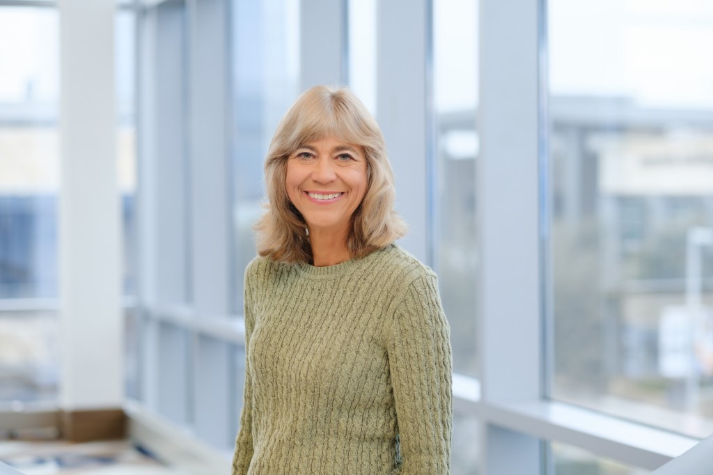 Woman smiling in a green sweater, standing near large windows with natural light. omni dallas headshot photographer nicole caldwell