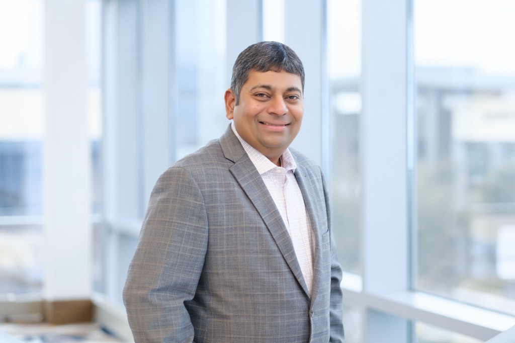 An individual in a gray suit stands confidently beside large windows with a bright, modern interior backdrop. omni dallas headshot photographer nicole caldwell