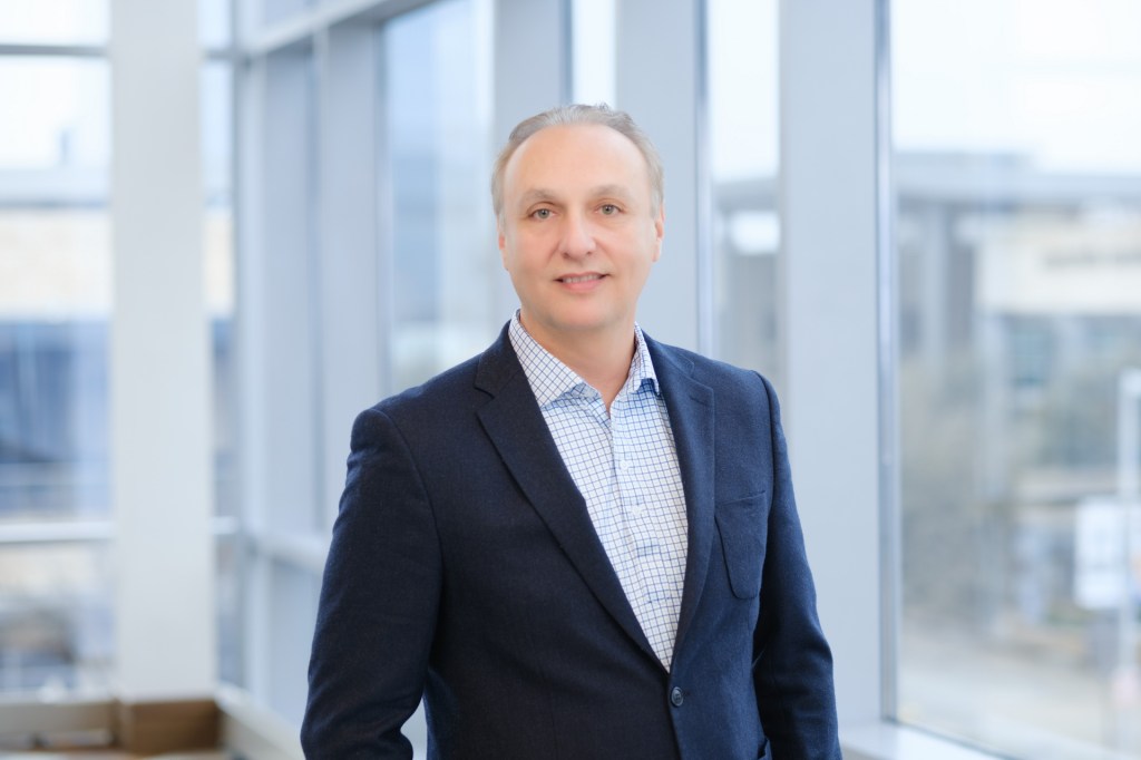 A professional man in a dark blazer and patterned shirt stands in front of a large window, with a bright and modern office environment in the background. omni dallas headshot photographer nicole caldwell