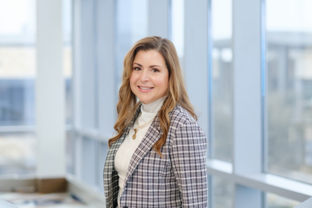 Professional headshot of a woman smiling, wearing a plaid blazer and white turtleneck, standing in front of large windows with natural light. omni dallas headshot photographer nicole caldwell