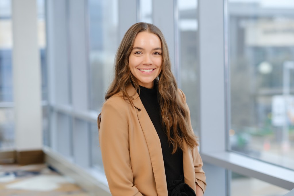 A young woman with long, wavy hair smiles at the camera while standing in front of a large window. She wears a beige blazer over a black top, with a blurred city view visible outside. omni dallas headshot photographer nicole caldwell