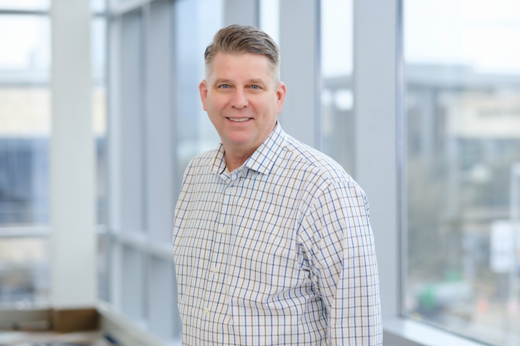 A smiling man in a checked shirt stands in front of large windows, showcasing an indoor setting with natural light. omni dallas headshot photographer nicole caldwell