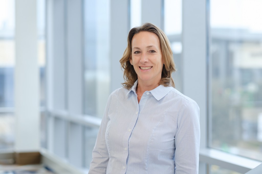 A smiling woman with shoulder-length hair stands in a well-lit indoor space, wearing a light blue button-up shirt. omni dallas headshot photographer nicole caldwell