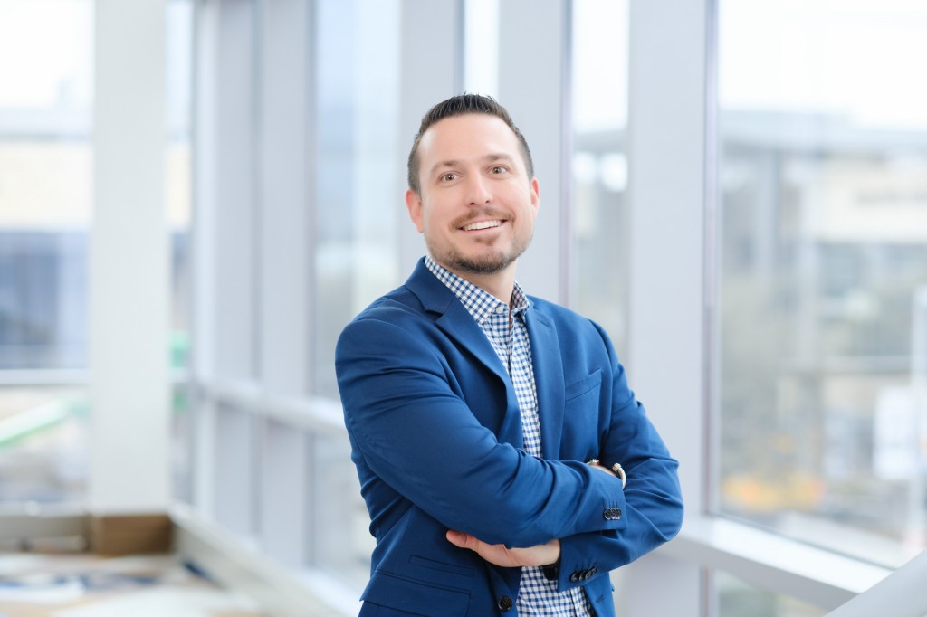 A professional man smiling confidently while standing next to large windows in a well-lit environment. omni dallas headshot photographer nicole caldwell
