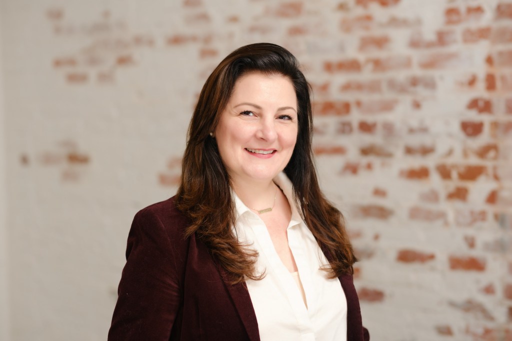 A woman with long, brown hair smiles at the camera while wearing a white blouse and a dark blazer. The background features a textured wall with a mix of white and red brick. Orange County Photo studio for headshots nicole caldwell