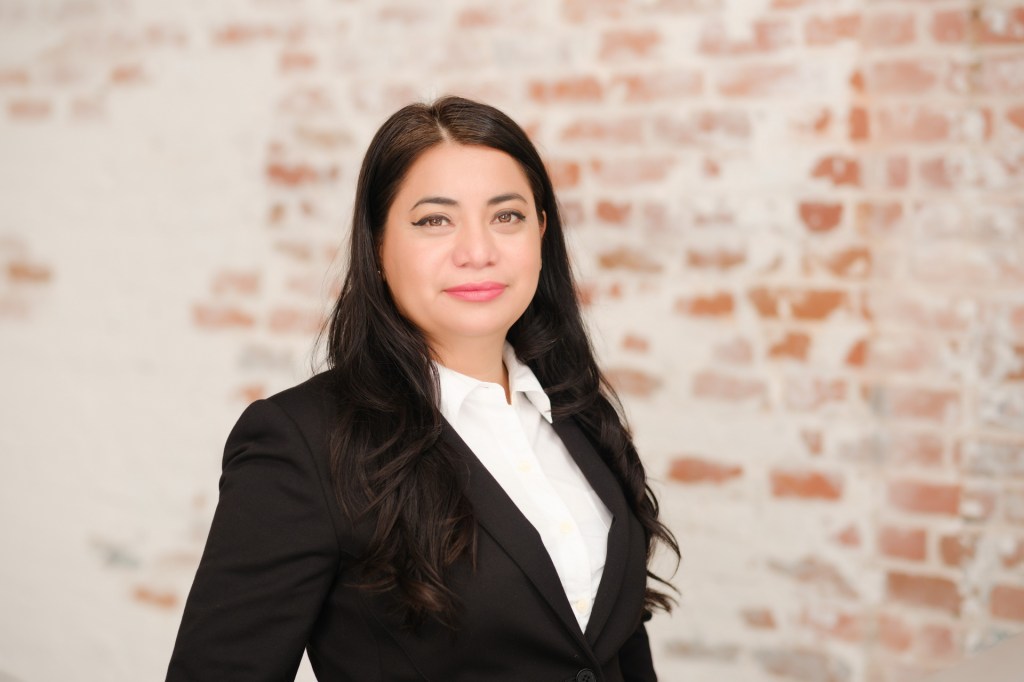 A woman with long black hair poses confidently in a black suit and white shirt against a brick wall background. Orange County Photo studio for headshots nicole caldwell