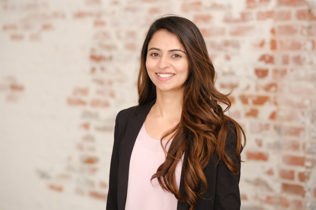 A smiling woman with long, wavy hair wearing a black blazer over a pink blouse, standing in front of a brick wall. Orange County Photo studio for headshots nicole caldwell
