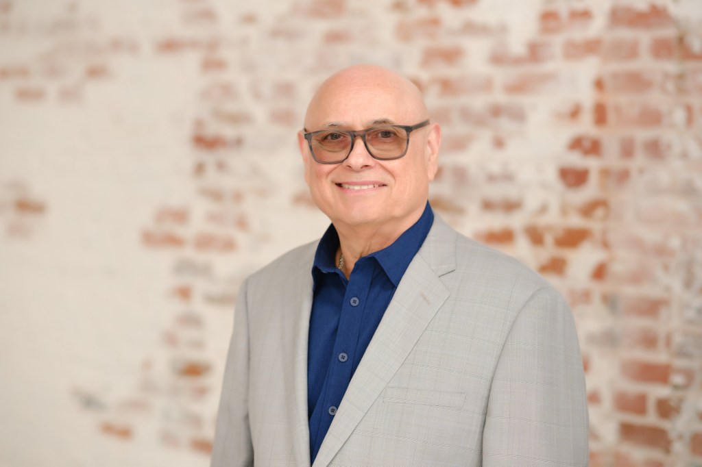 A man with bald head wearing glasses and a light gray suit, smiling in front of a brick wall. Orange County Photo studio for headshots nicole caldwell