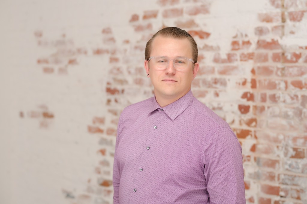 A man with glasses wearing a purple shirt stands in front of a brick wall. Orange County Photo studio for headshots nicole caldwell