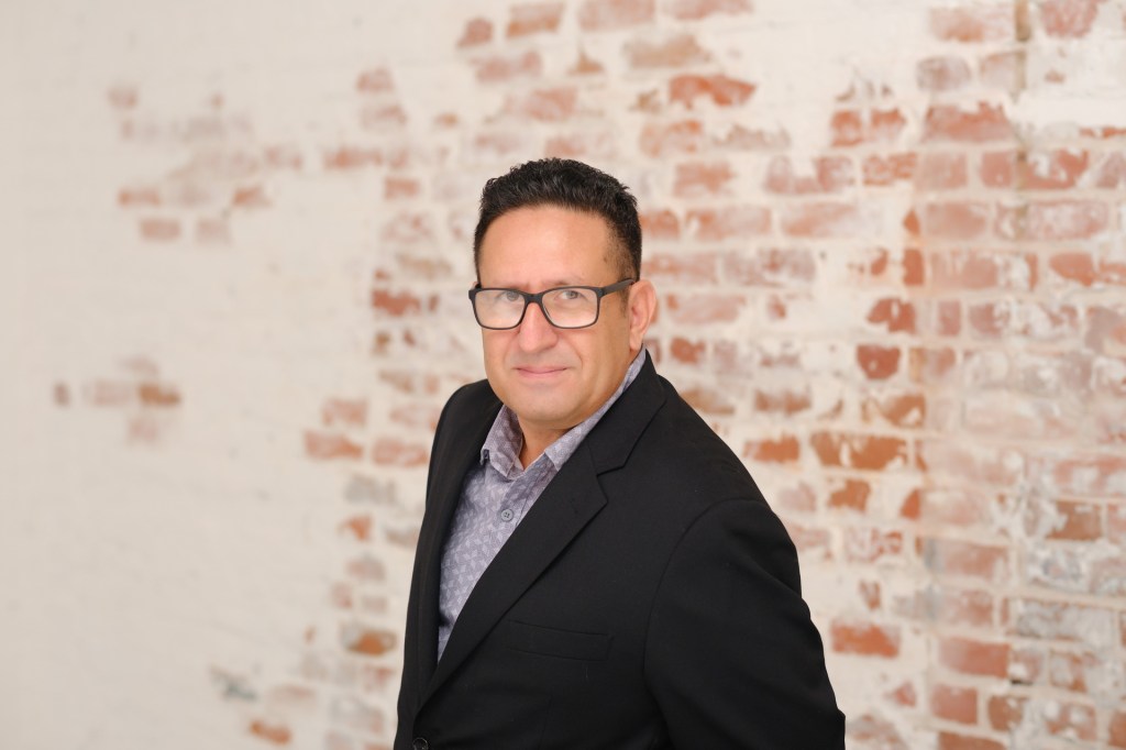 A man in a black blazer and glasses is standing against a textured brick wall, smiling slightly and looking directly at the camera. Orange County Photo studio for headshots nicole caldwell