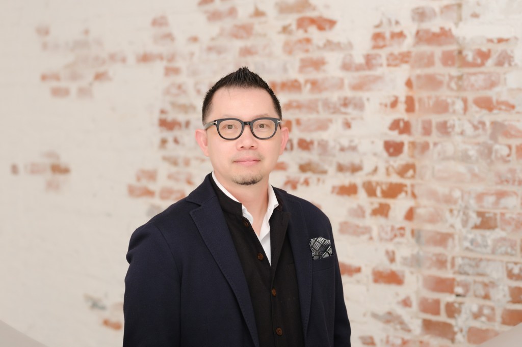 A person wearing glasses and a dark blazer stands against a white-brick wall with a slight smile. Orange County Photo studio for headshots nicole caldwell