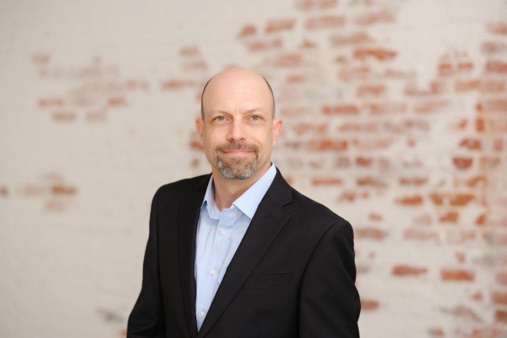 A man in a black suit and light blue shirt stands against a textured brick wall, smiling at the camera. Orange County Photo studio for headshots nicole caldwell