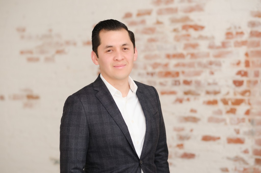 A man in formal attire, wearing a navy blazer over a white shirt, stands in front of a rustic brick wall, smiling confidently. Orange County Photo studio for headshots nicole caldwell