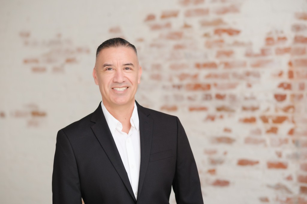 A professional man smiling, wearing a black suit and white shirt, posed in front of a textured brick wall. Orange County Photo studio for headshots nicole caldwell